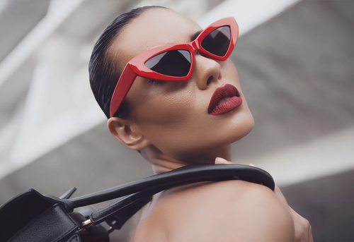 A woman wearing red cat-eye sunglasses and bold red lipstick looks over her shoulder, holding a black bag against a blurred, modern architectural background.